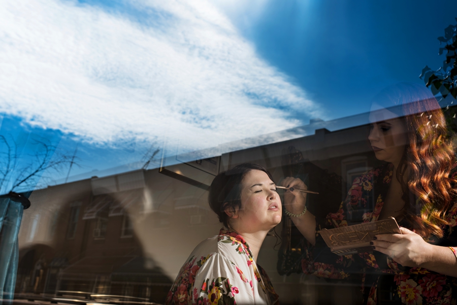 A bride getting her makeup done with the window reflecting the blue sky above.